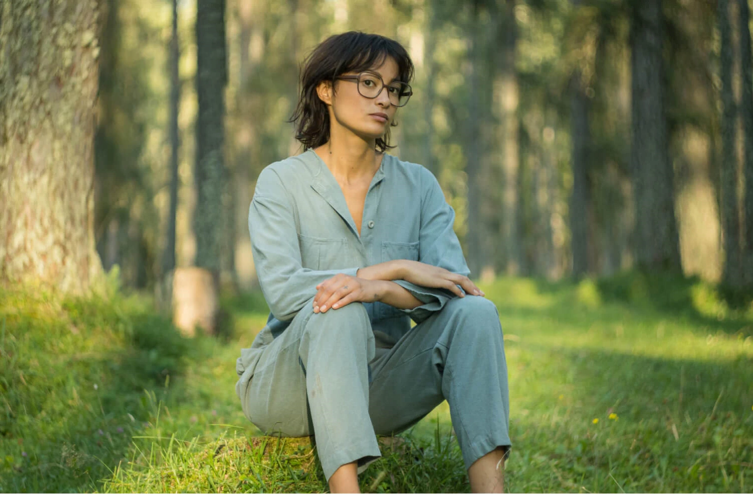 Femme en combinaison vert sauge assise dans une forêt, portant des lunettes rondes Rolf